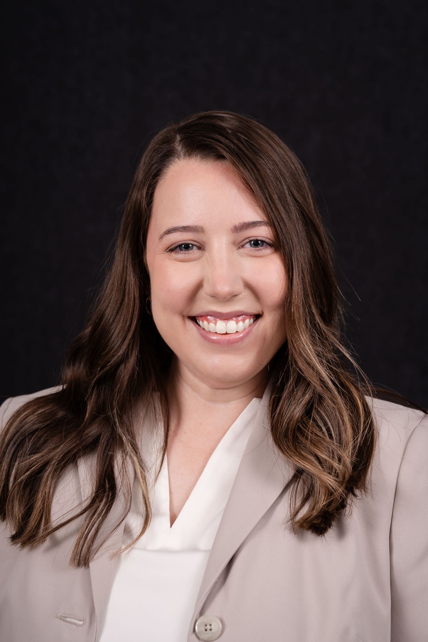 Professional headshot portrait with natural smile wearing beige blazer and white blouse against dark background.