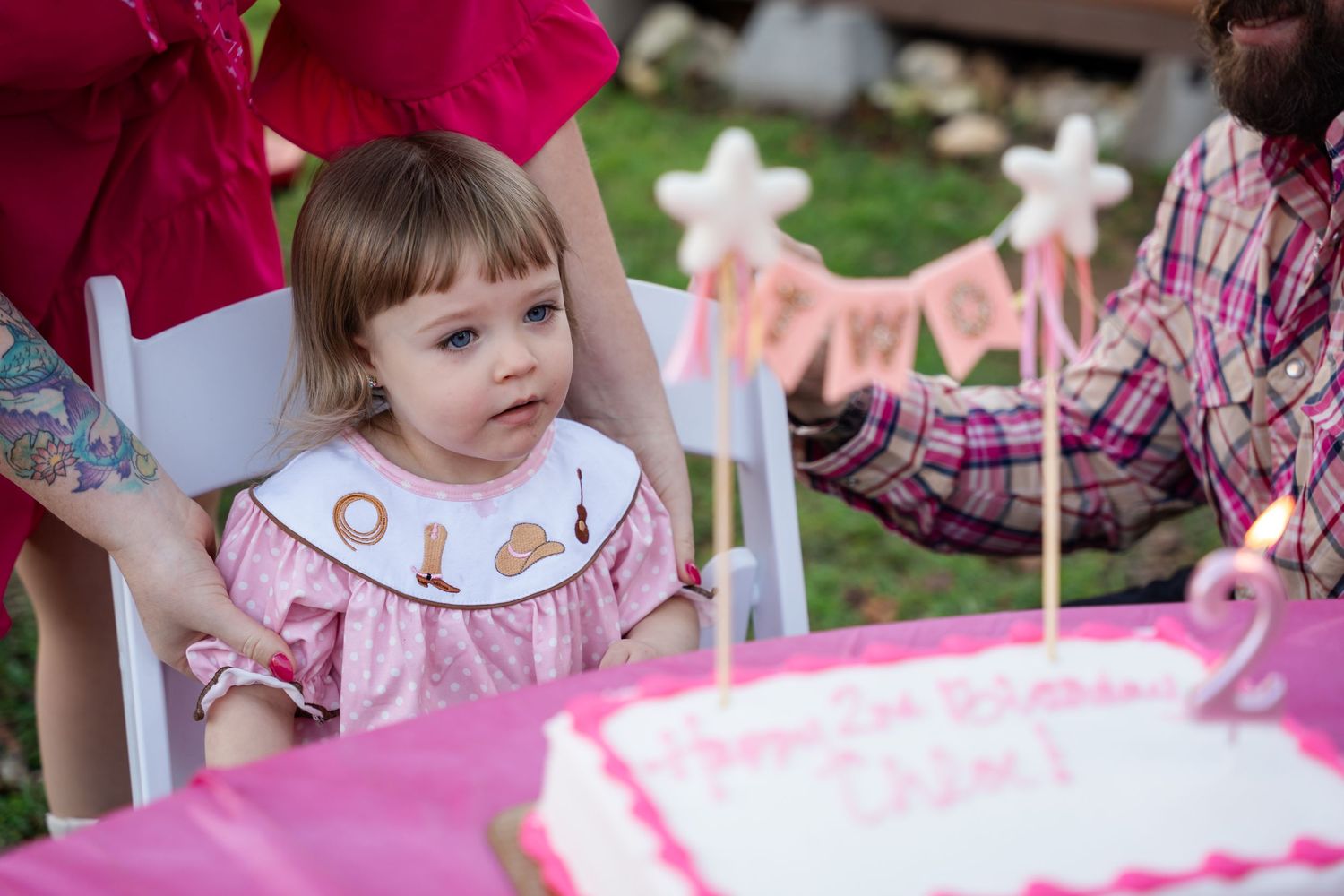 Young child in pink dress sitting at birthday party table with lit candles on cake.