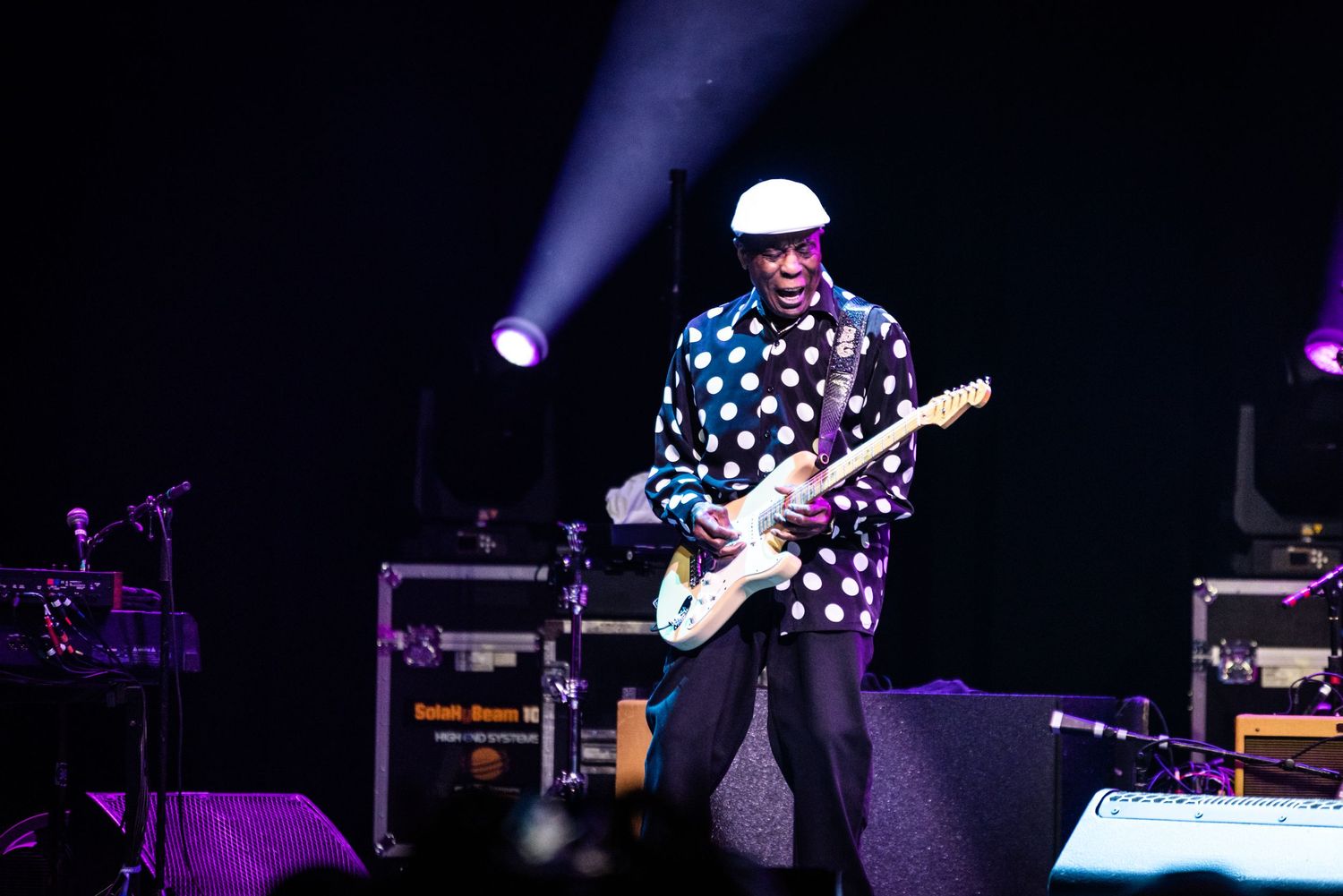 Musician in polka dot shirt plays guitar on dark stage with purple lighting effects.