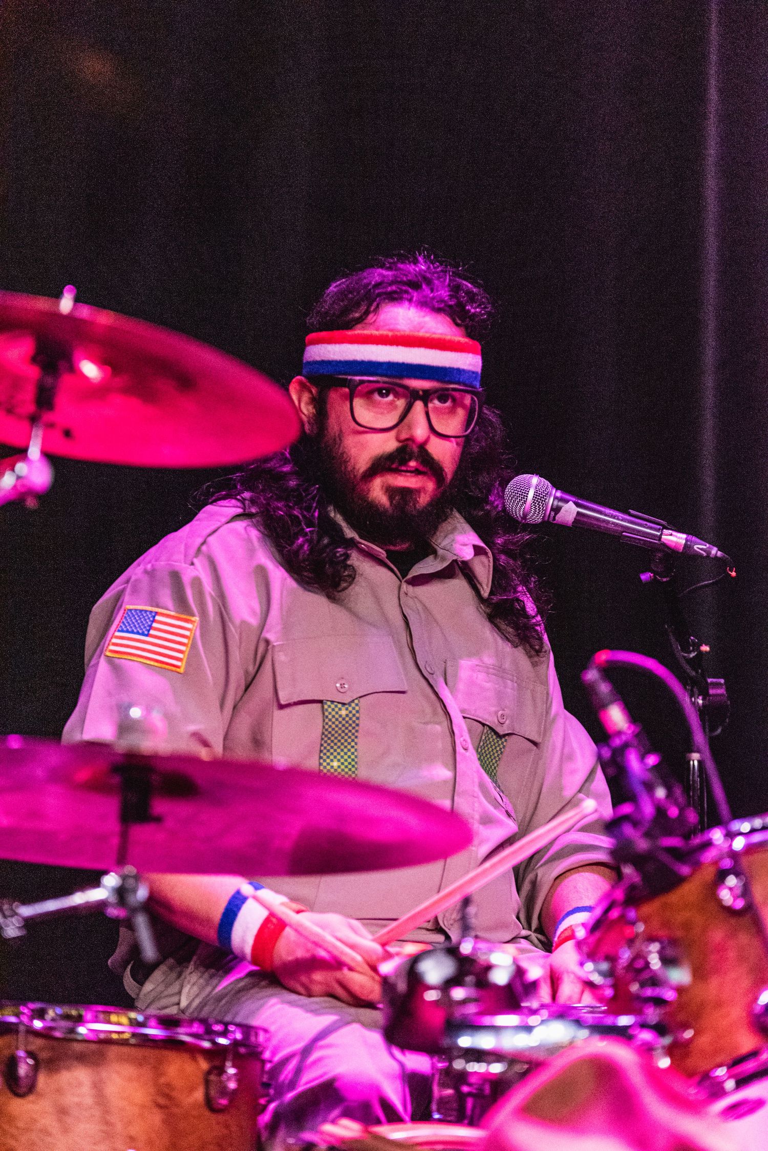 Drummer in scout-style uniform with glasses performs under purple stage lighting.