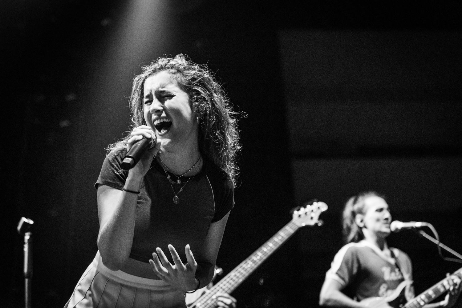 Concert performer sings into microphone with band member visible in background on dimly lit stage.