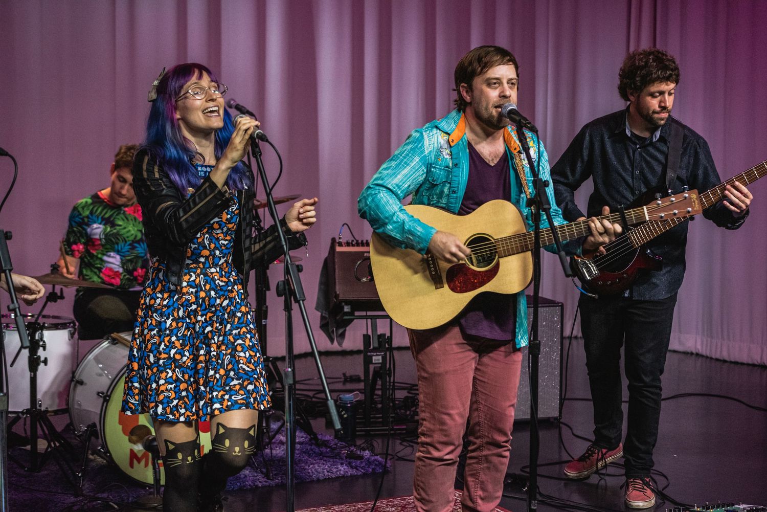 Band performs on stage with singer and guitarists against purple backdrop.