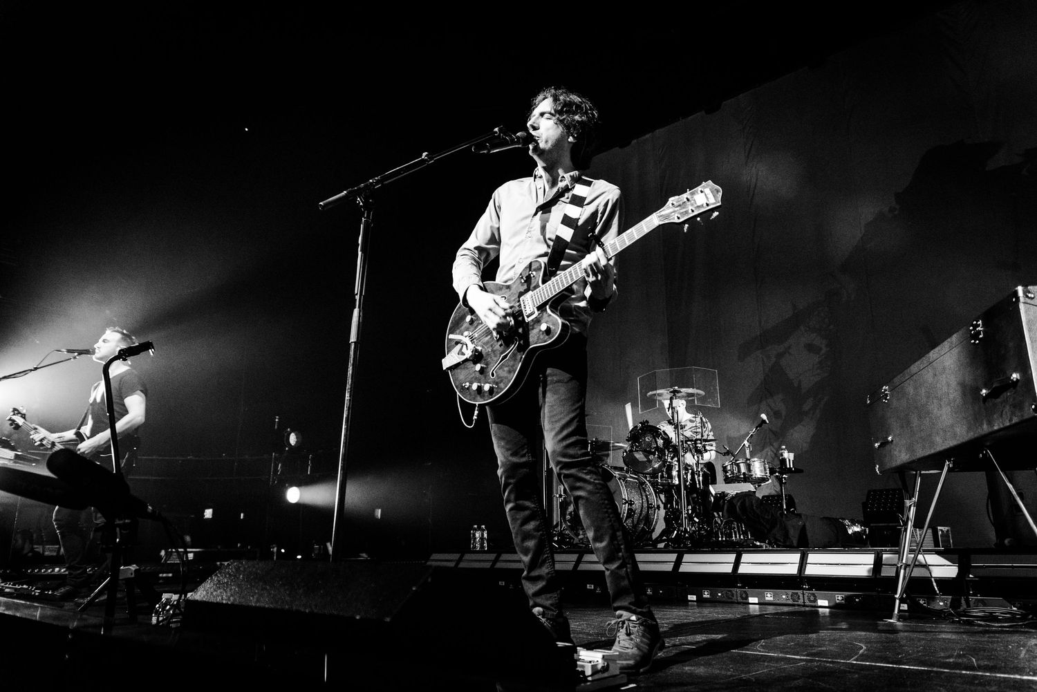 Black and white wide shot of band performing on stage with dramatic concert lighting.
