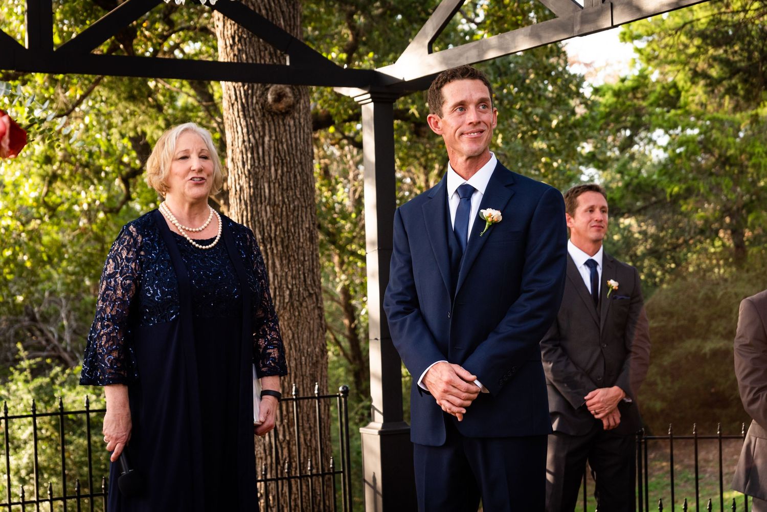 A formal outdoor wedding ceremony with attendees standing beneath trees.