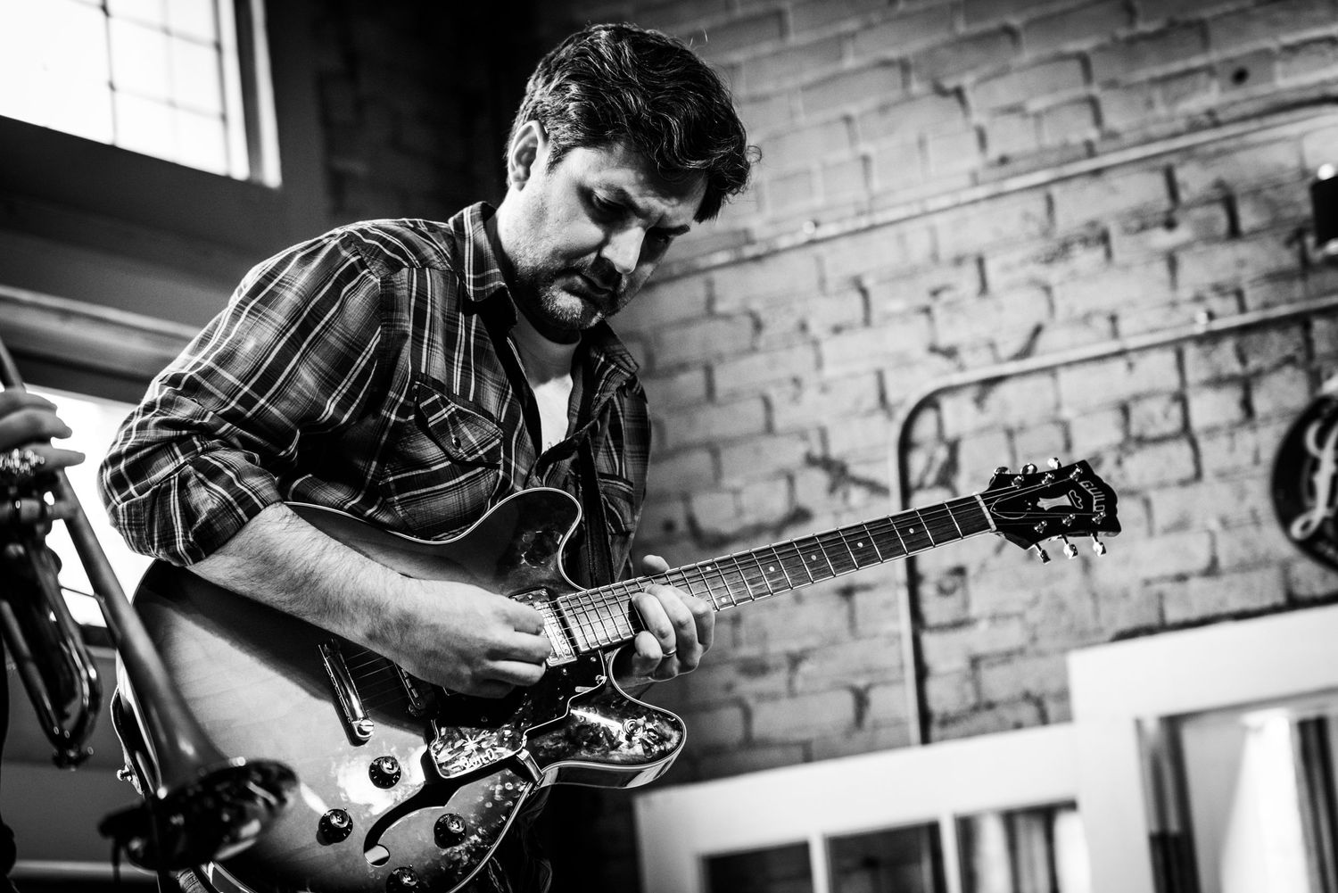 Black and white photograph of guitarist playing electric guitar against brick wall background.
