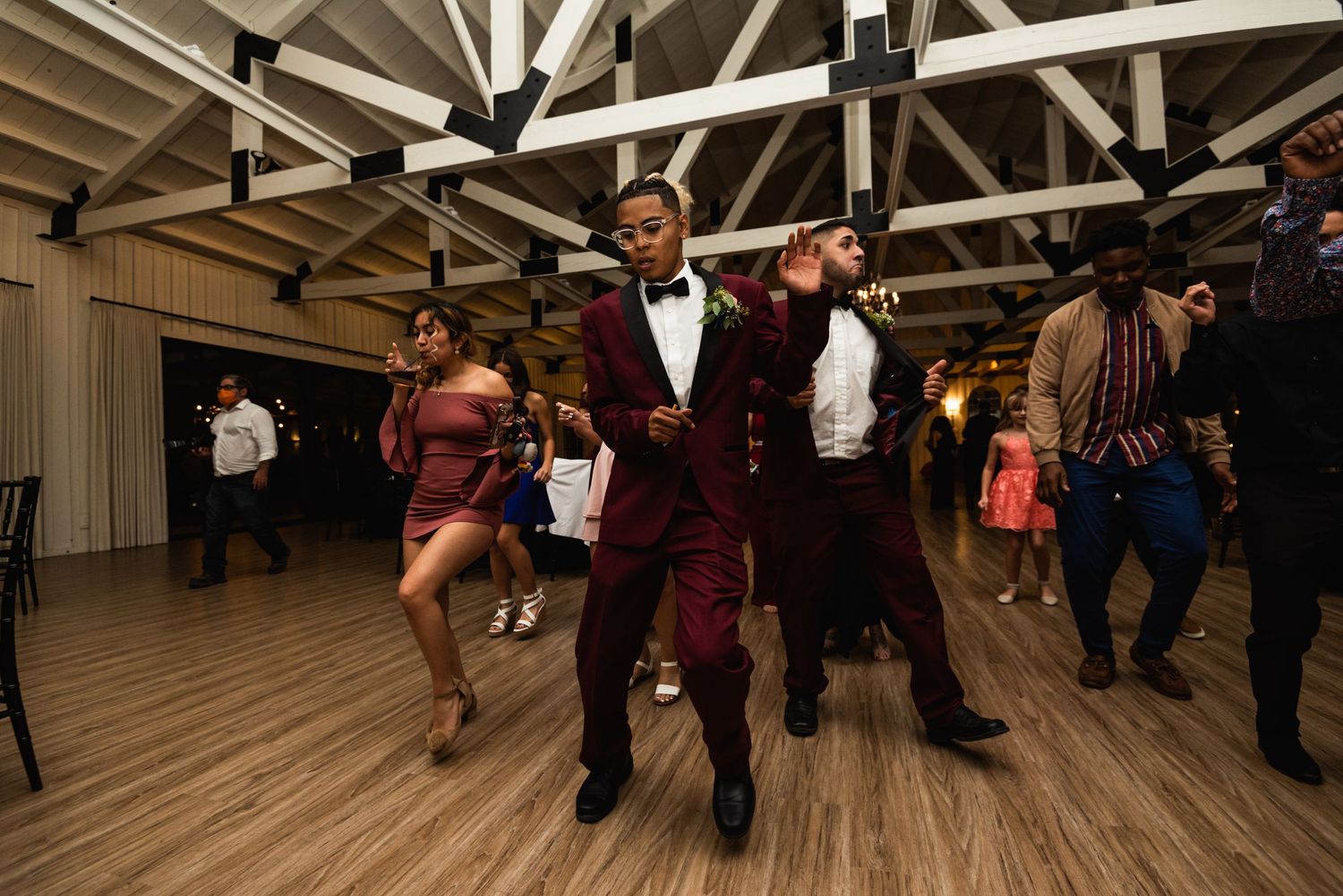 Wedding guests energetically dancing on a wooden dance floor at an indoor reception venue.