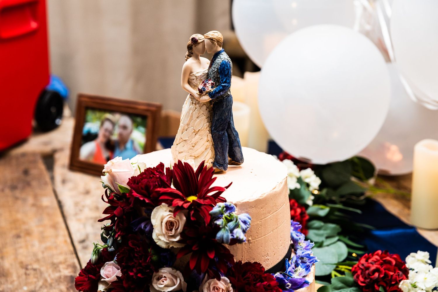 Wedding cake topped with traditional figurines features deep red and burgundy floral decorations.