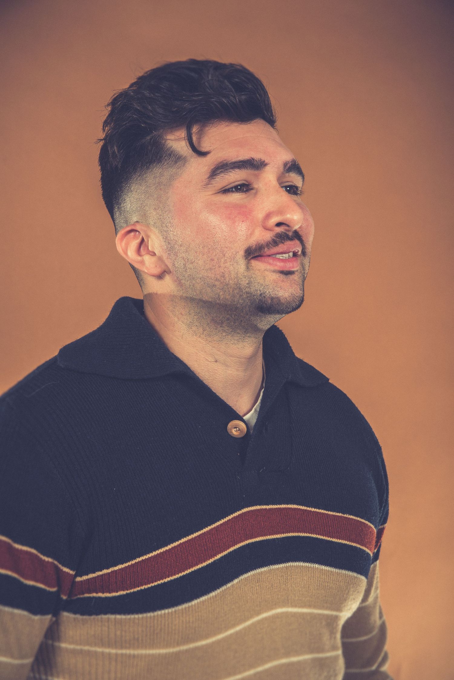 Portrait of someone in striped sweater against orange backdrop with styled hair.