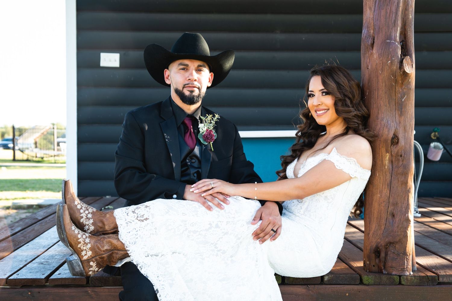 Newlyweds in western attire sitting on rustic wooden bench.