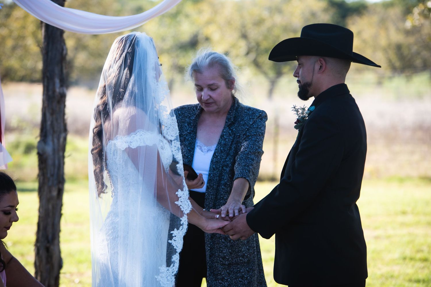 Wedding ceremony takes place under trees with an officiant and couple at a rustic outdoor venue.