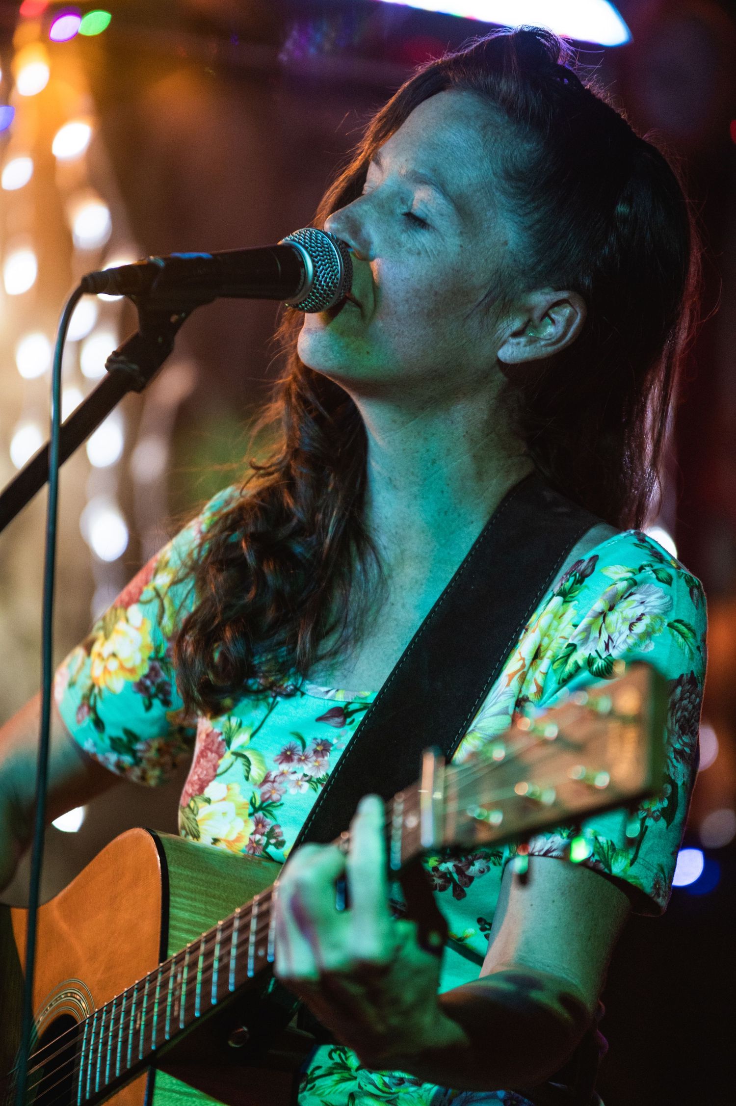 Singer-songwriter performs acoustic guitar under blue-green stage lighting in floral dress.