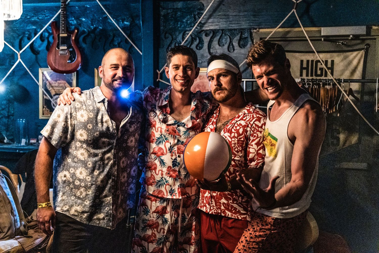 Band members in colorful shirts posing together in front of blue industrial backdrop.