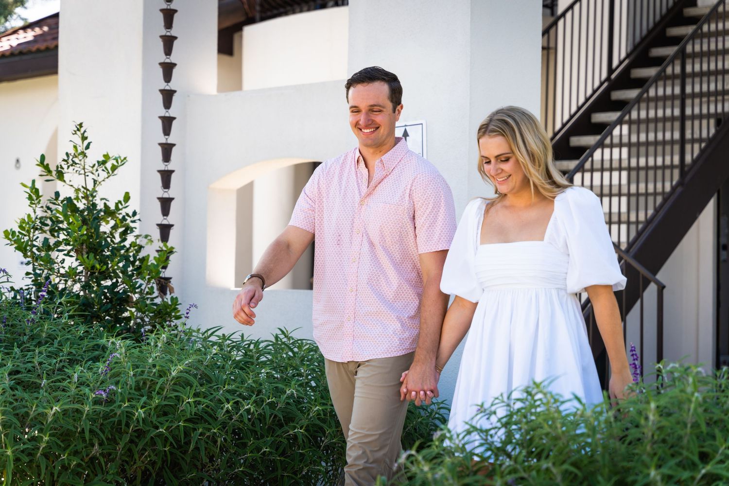 A couple takes a romantic stroll through a garden path beside a white building with stairs.