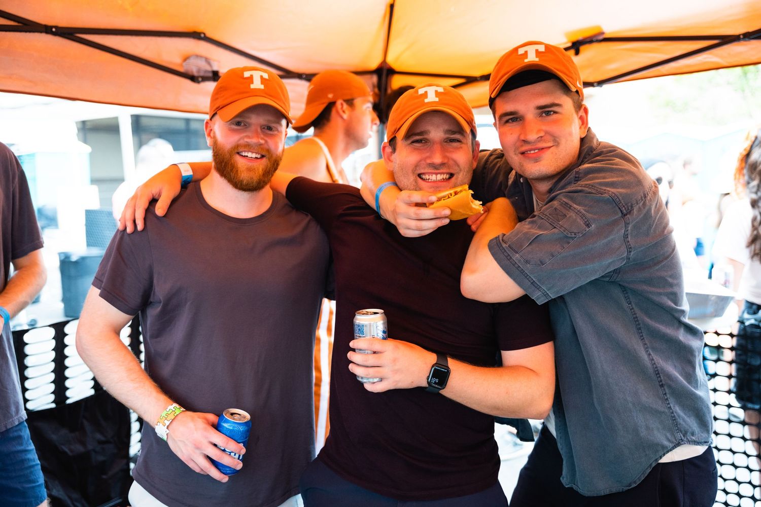Three friends in casual wear sharing drinks under orange tent at outdoor party.