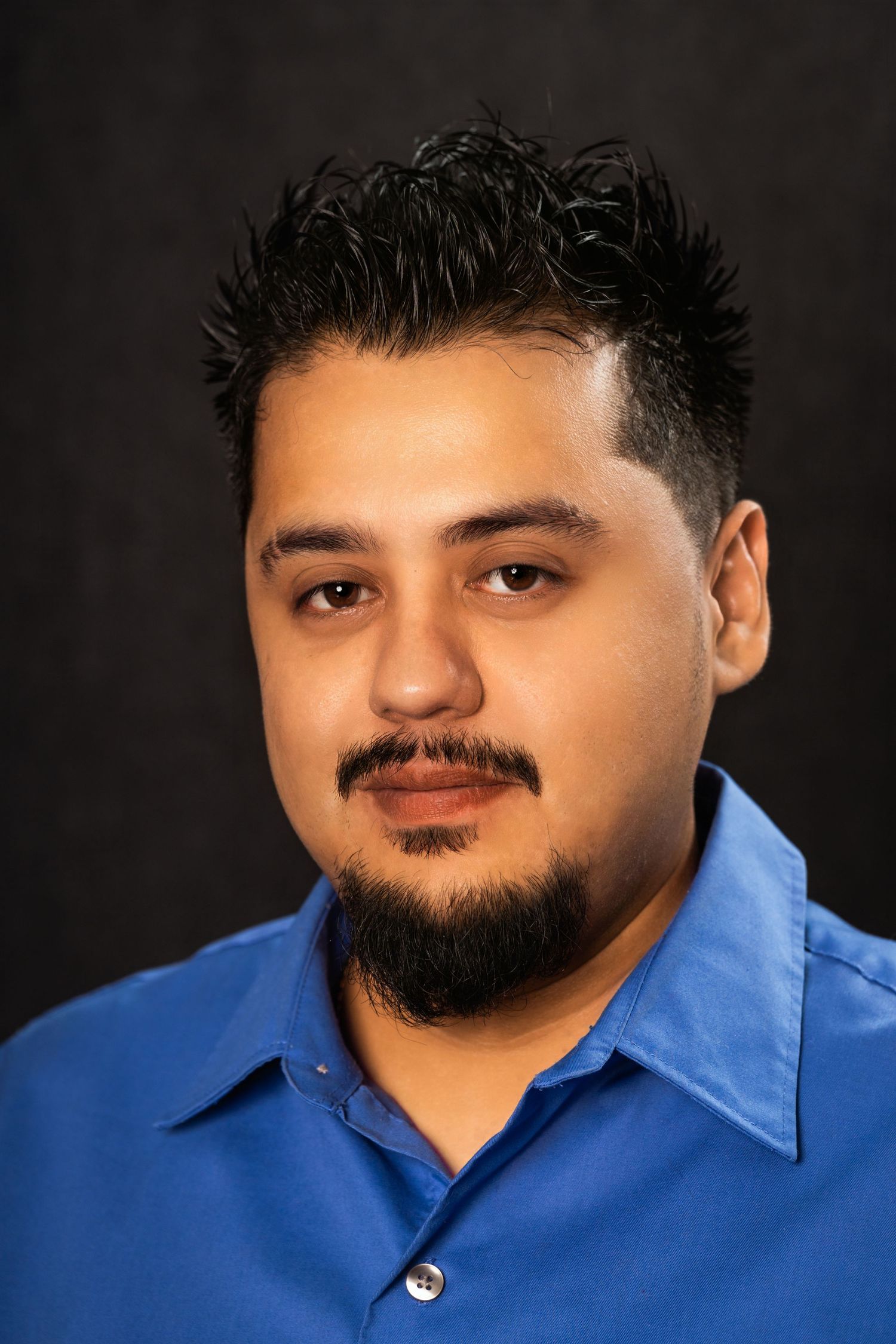 Close up studio portrait of a man in blue shirt with styled hair against dark background.