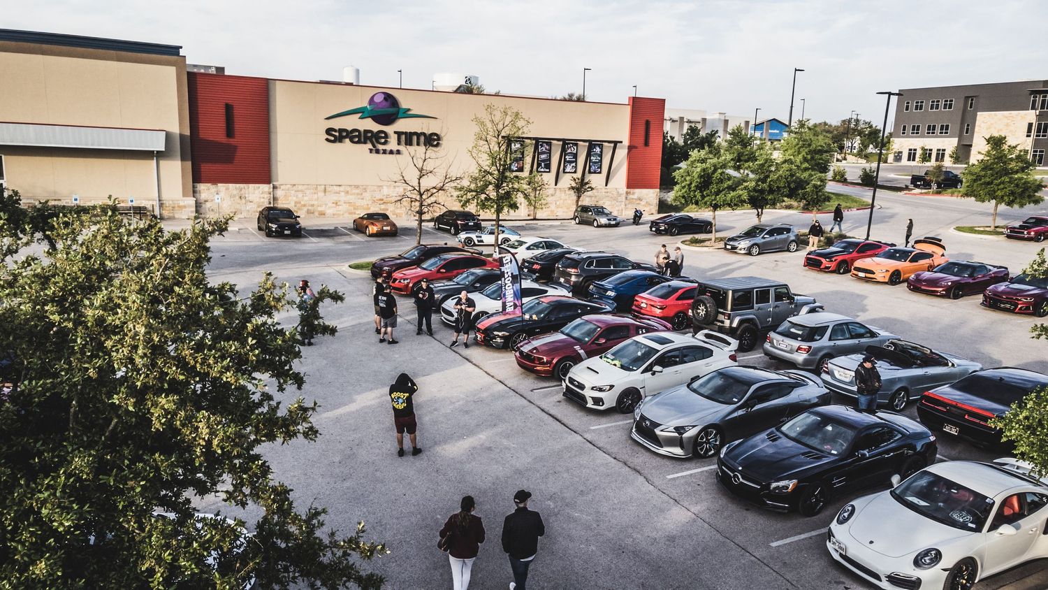 Bird's eye view of cars gathered in a commercial parking lot for an automotive event.