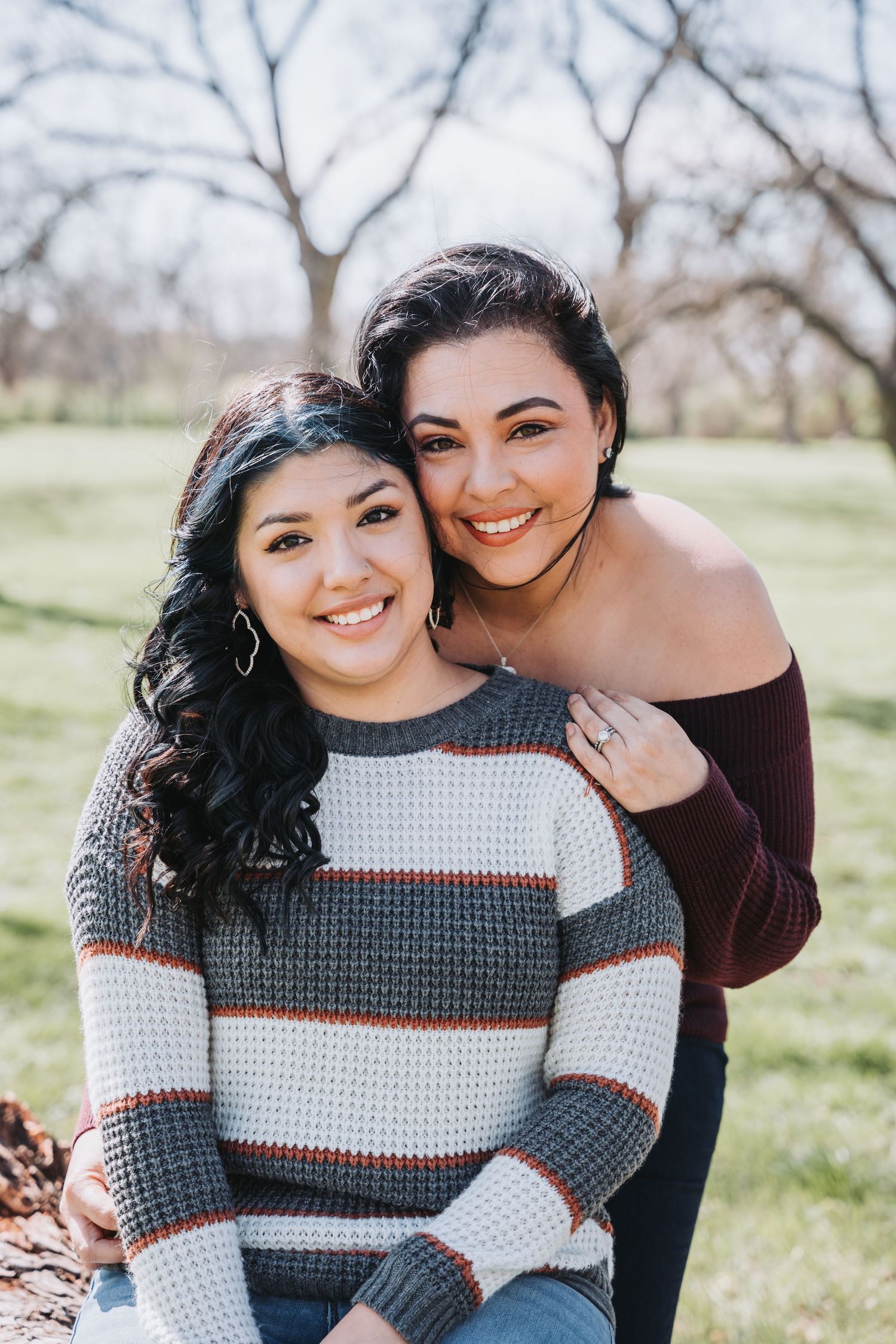 Two people embrace while smiling outdoors with trees and grass in background.