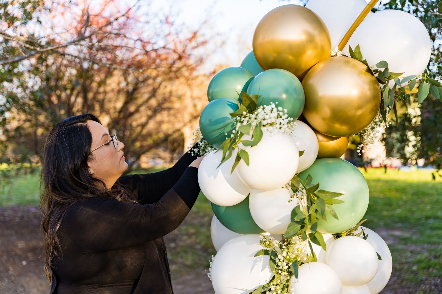 Person arranging a festive balloon arch with gold, mint, and white balloons outdoors.