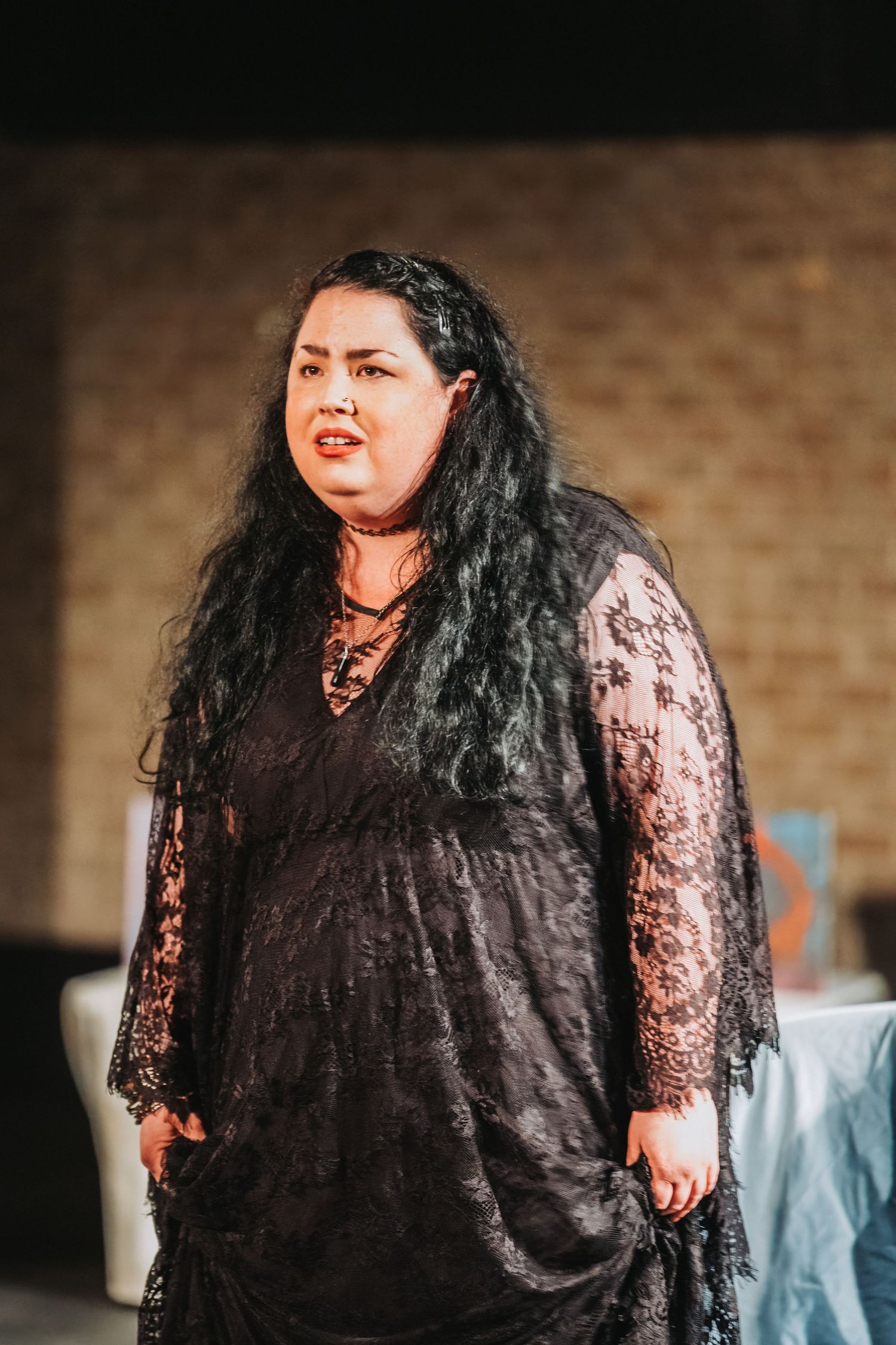 A performer in a black lace dress stands against a brick wall during a theatrical performance.
