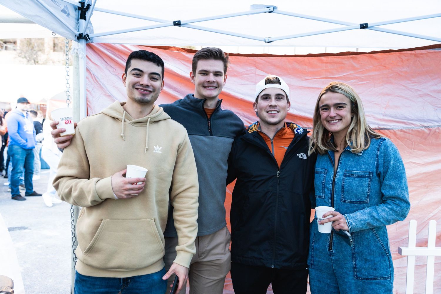 A group of friends pose together under a white tent at an outdoor event wearing casual attire.