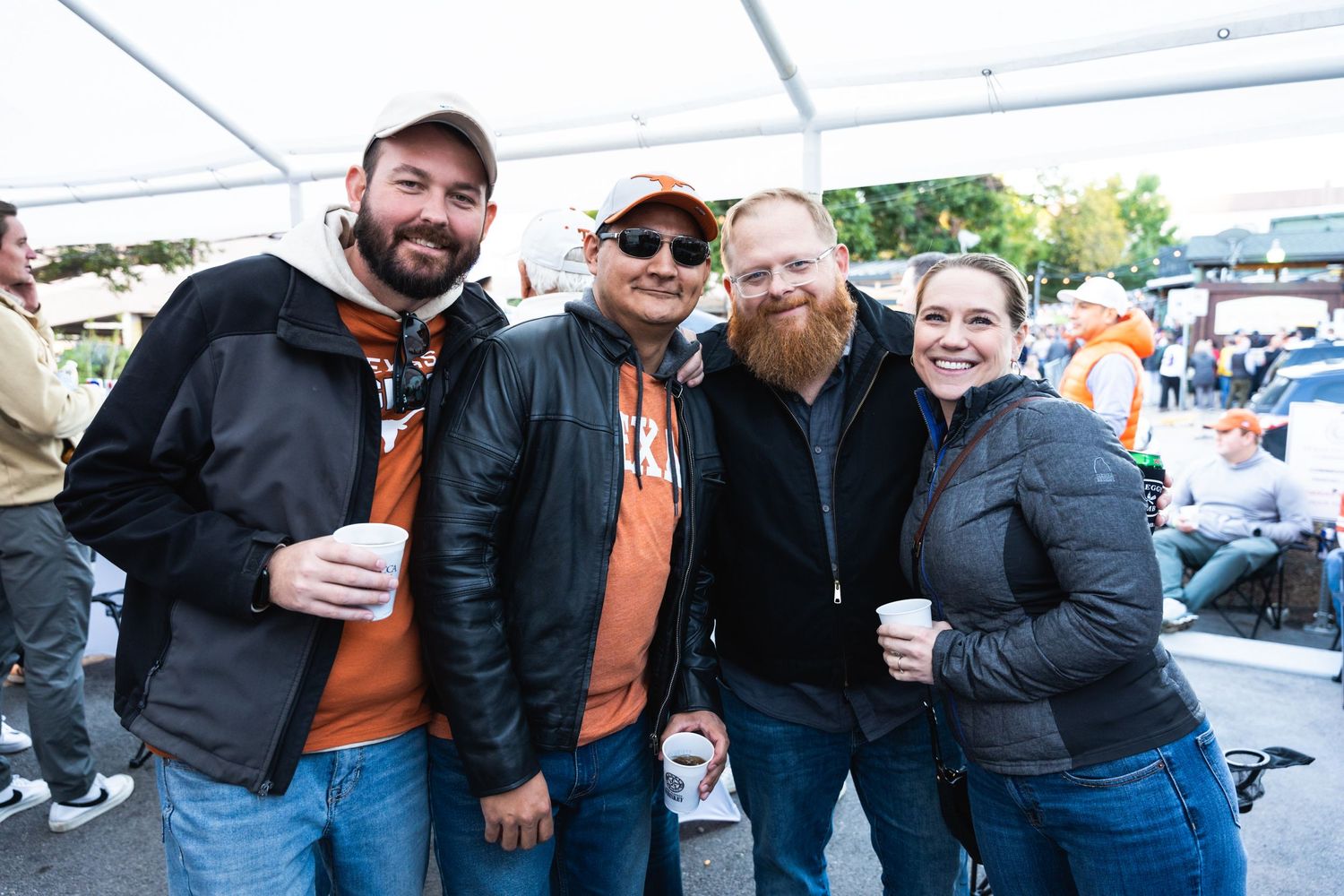 Four friends gather under a tent at an outdoor event wearing casual jackets and holding drinks.