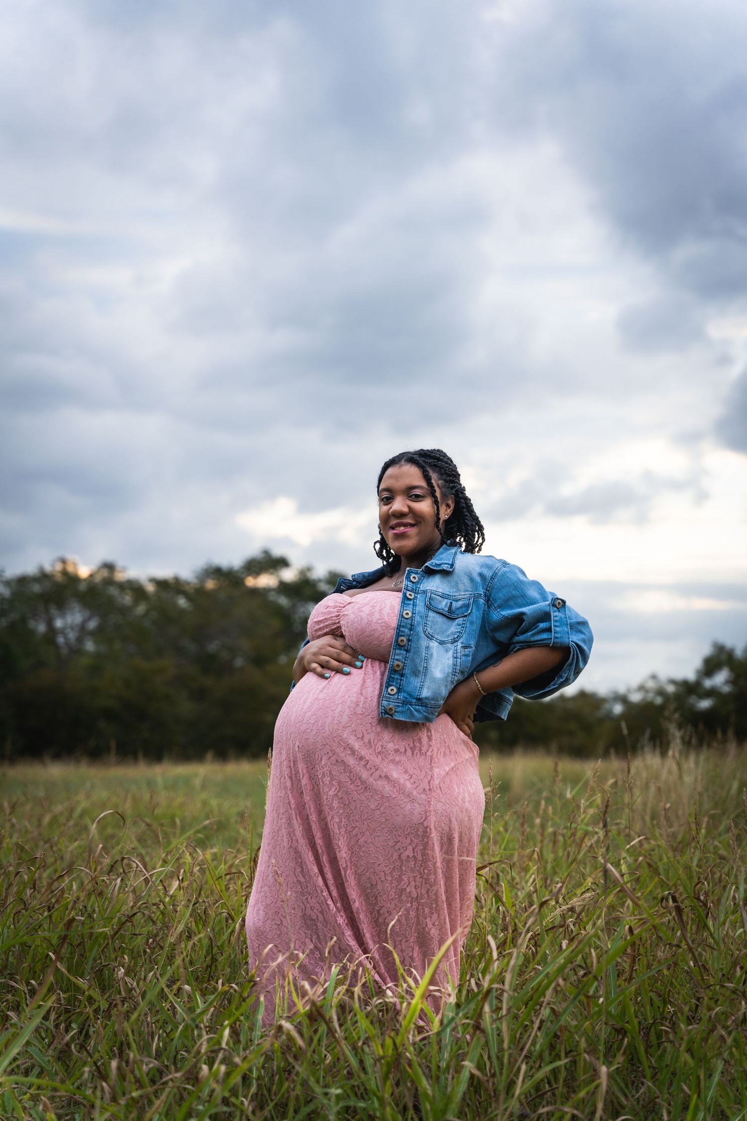 A maternity portrait in a field with dramatic cloudy sky featuring a pink dress and denim jacket.