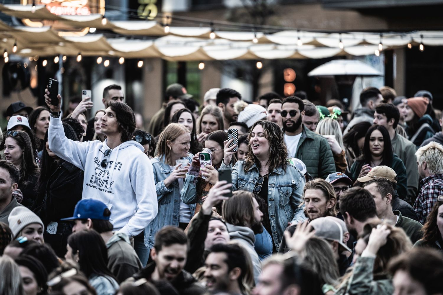 Large enthusiastic crowd gathering at an outdoor festival or concert with string lights overhead.