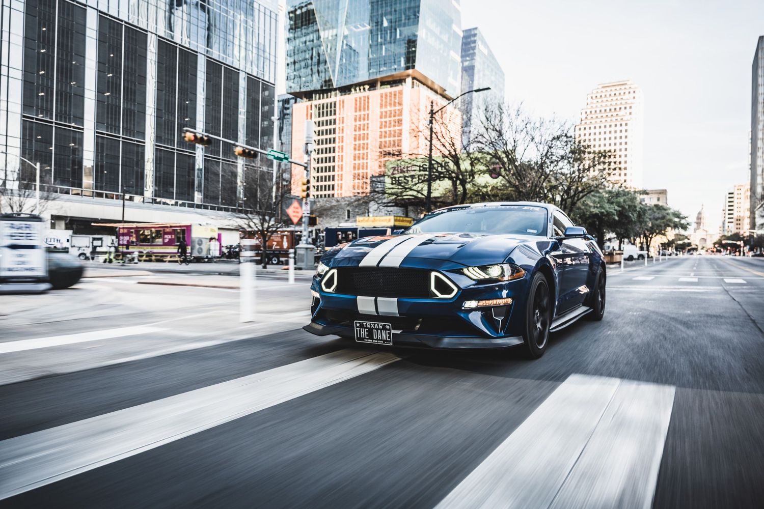 Modified blue Mustang GT driving through modern city intersection with glass skyscrapers.
