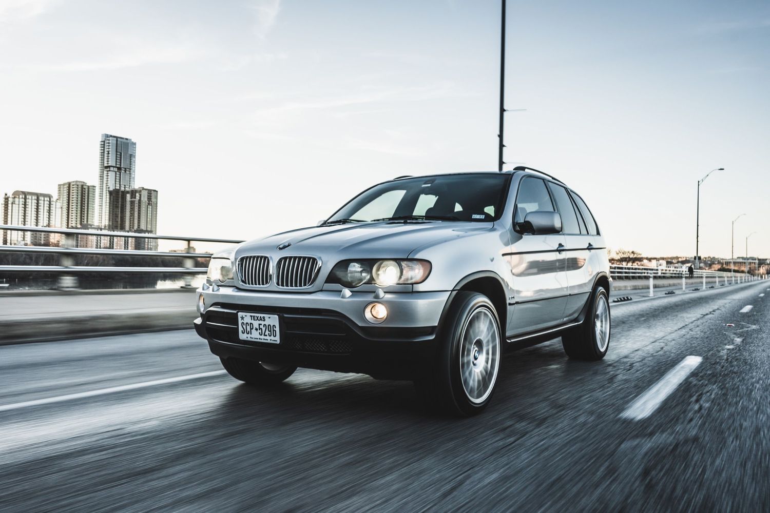 Silver BMW X5 SUV driving on highway with city skyline visible in distance.