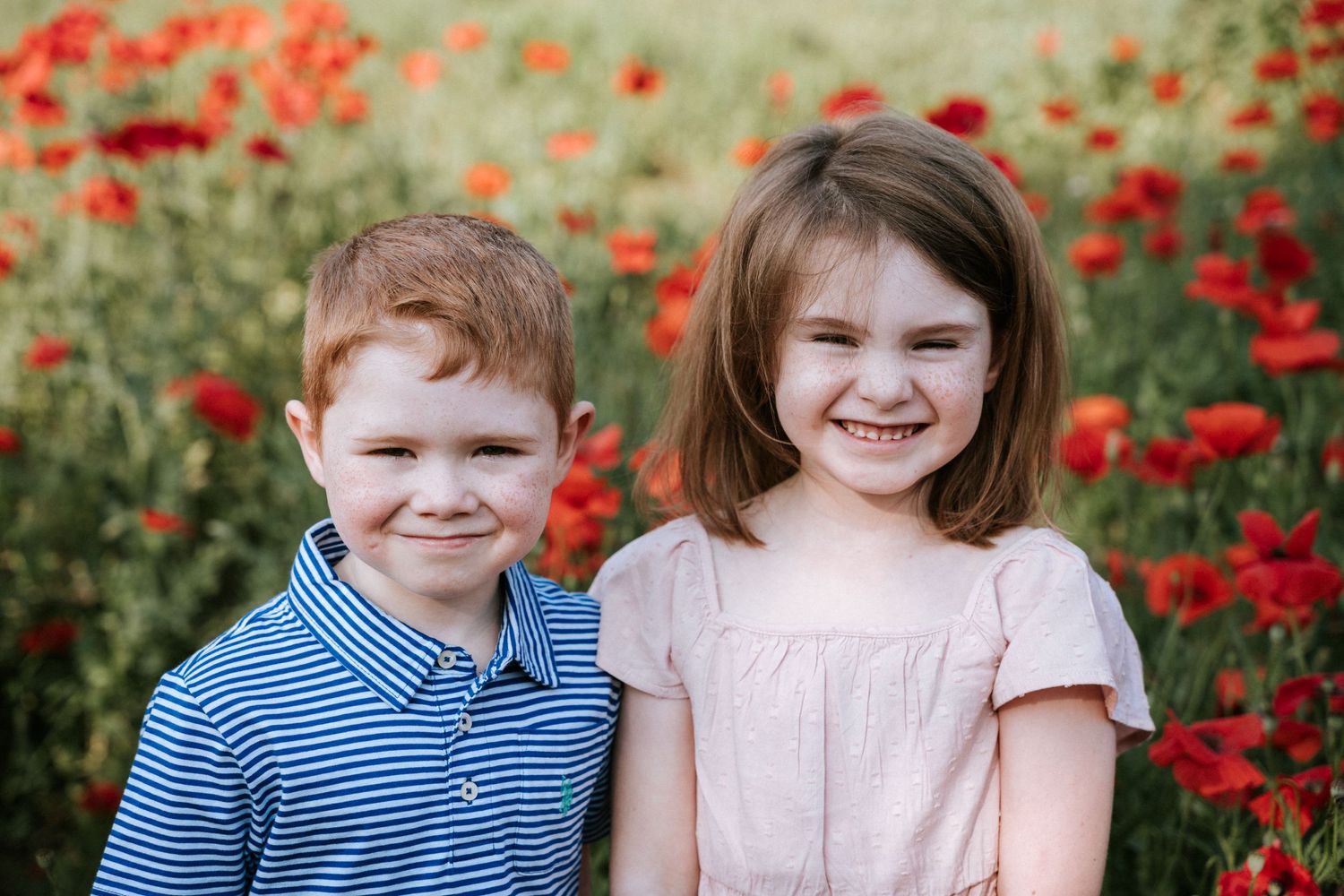 Two children smile at camera with vibrant red poppies in background.