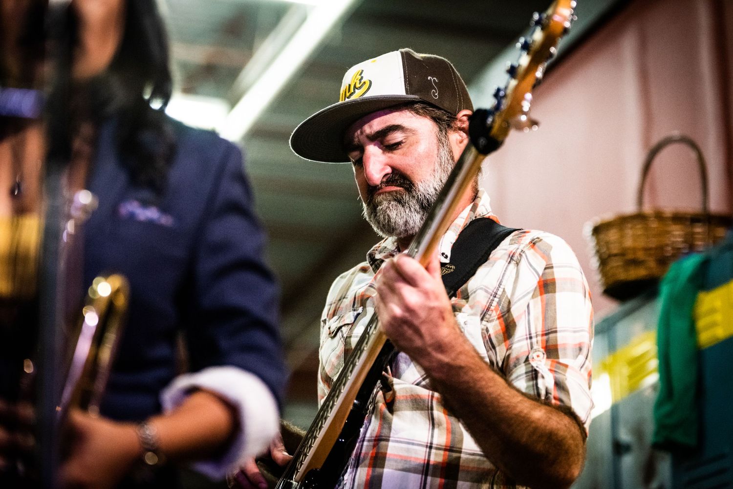 Guitarist in plaid shirt and cap performs with focused expression.