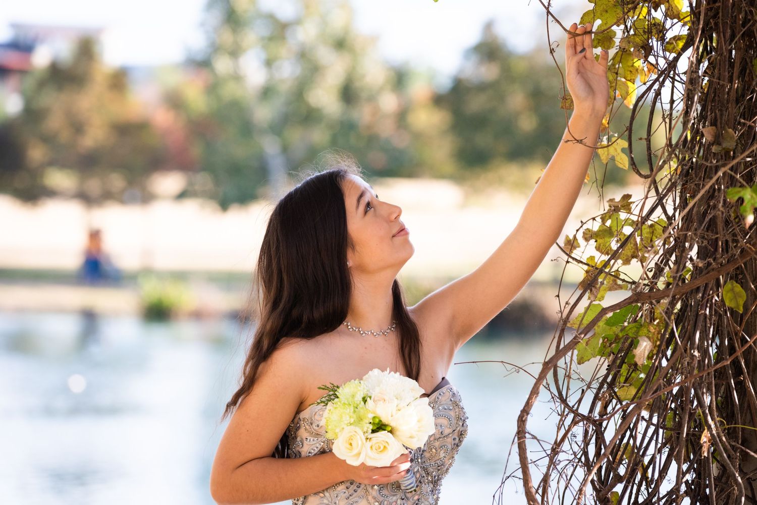 Bride in sparkly dress reaches up to touch tree branches by waterfront.