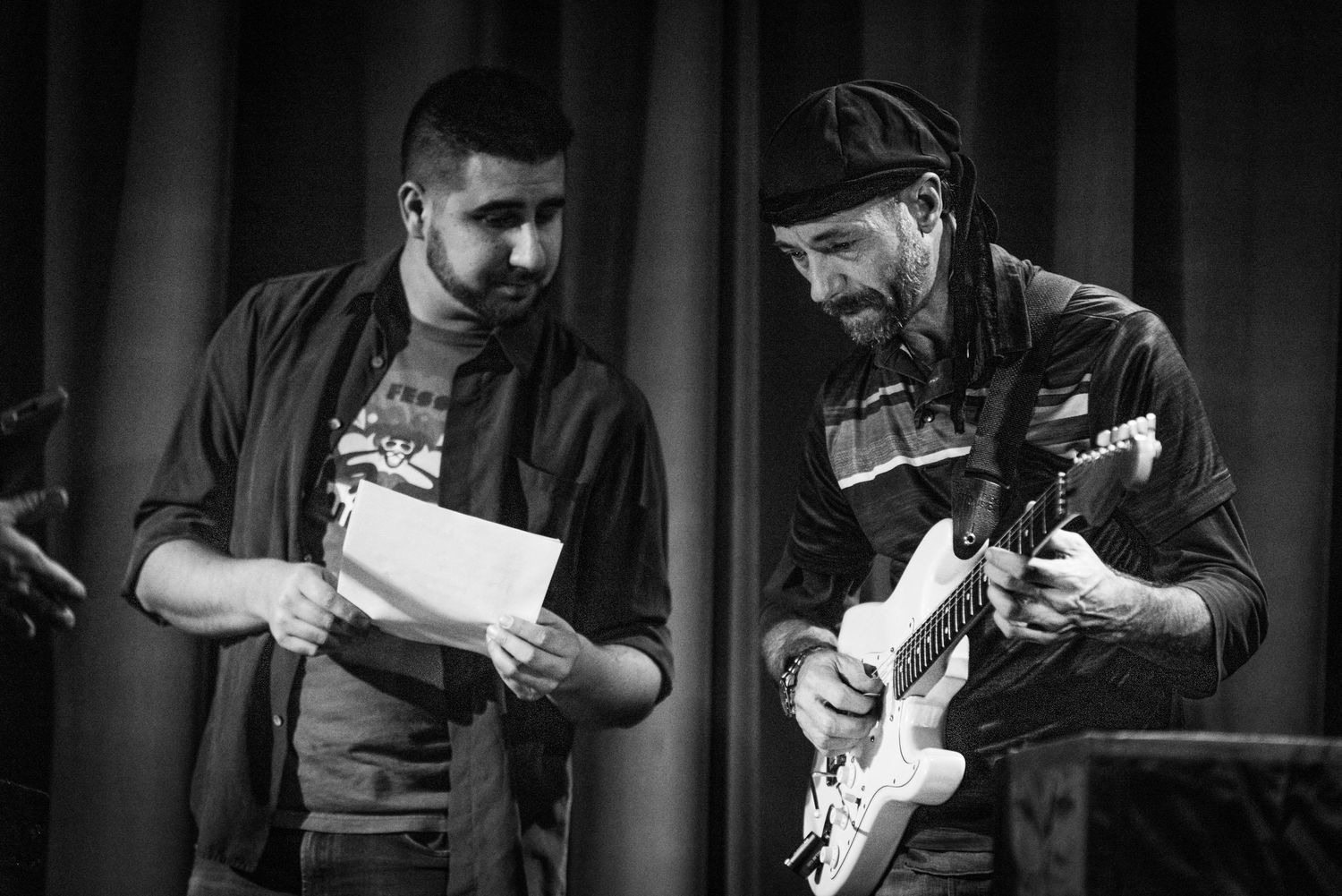 Two musicians collaborate on stage, one reading papers while another plays guitar in black and white.