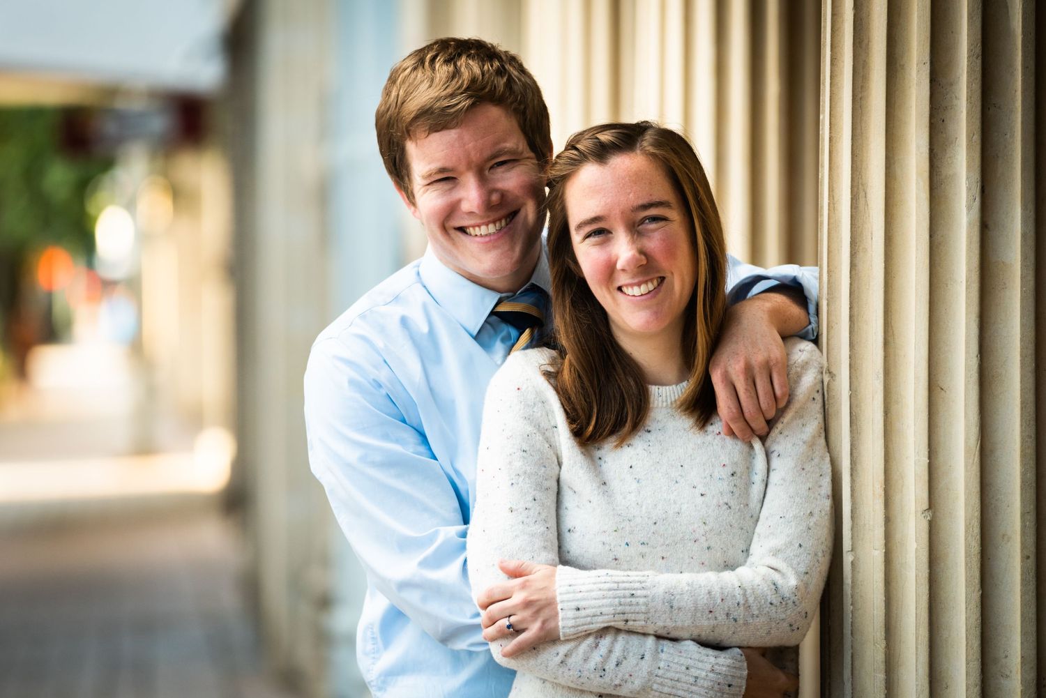 A couple poses together against a column in casual attire.