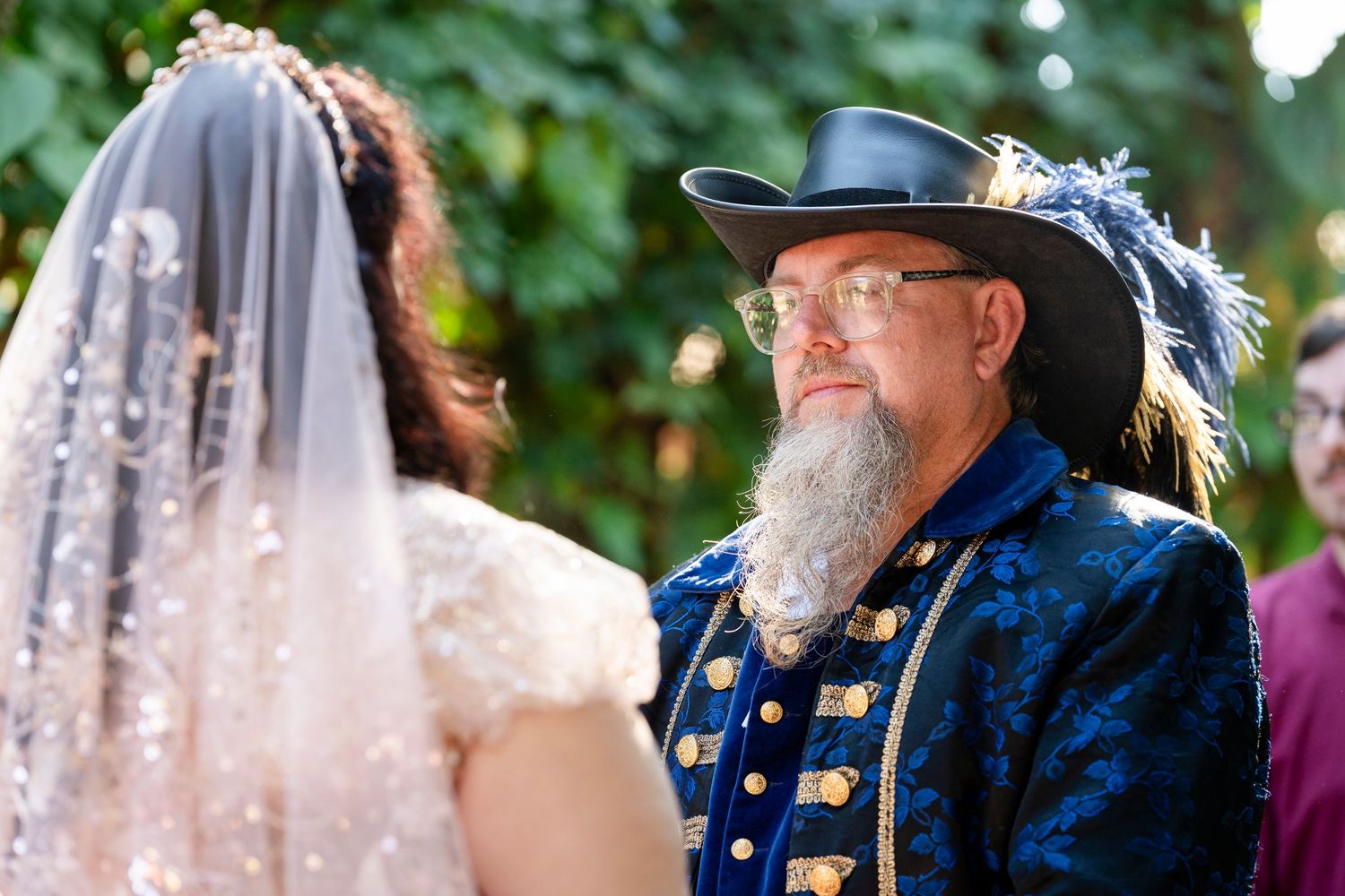 A wedding moment captured outdoors showing a person in an ornate blue colonial-style jacket with brass buttons and a feathered hat.
