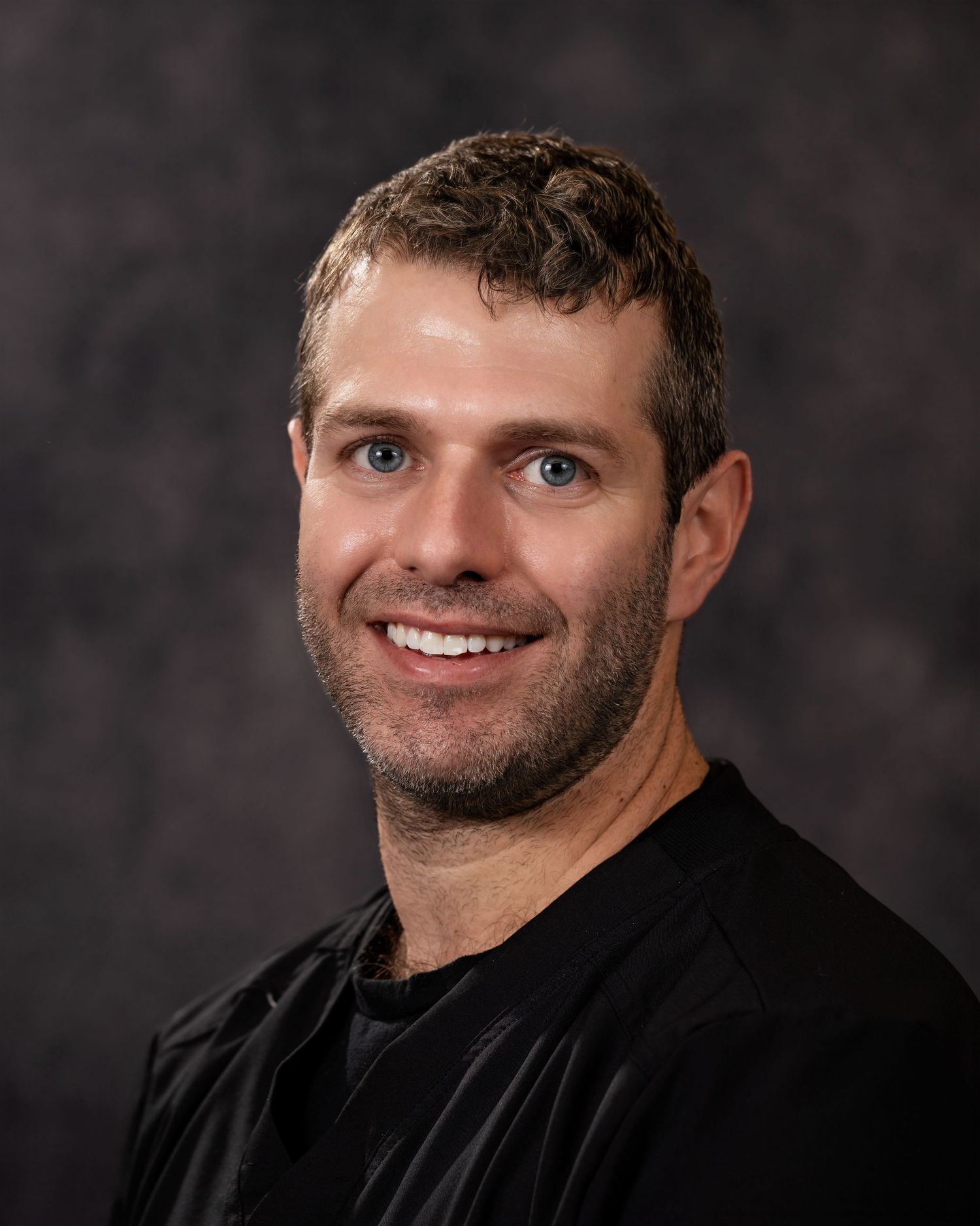 Professional headshot portrait of someone wearing a black shirt against a dark background with warm lighting.
