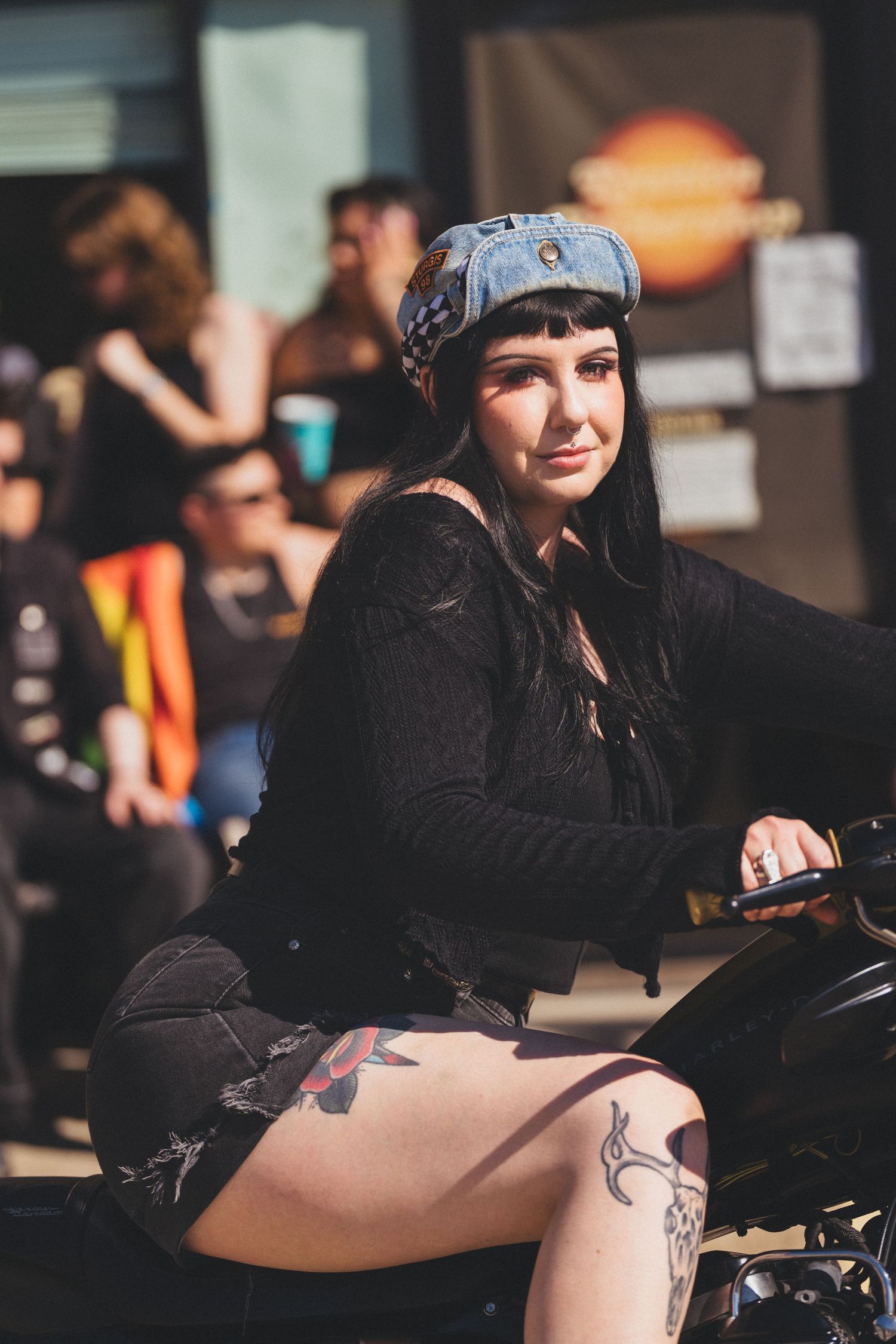 Biker wearing black clothing and bandana sits on motorcycle with visible leg tattoo in sunny outdoor setting.