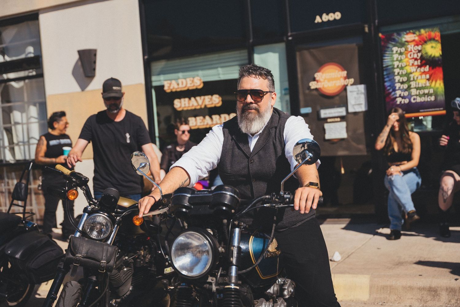 A group of motorcyclists gather outside storefronts on a sunny day during a motorcycle meetup event.