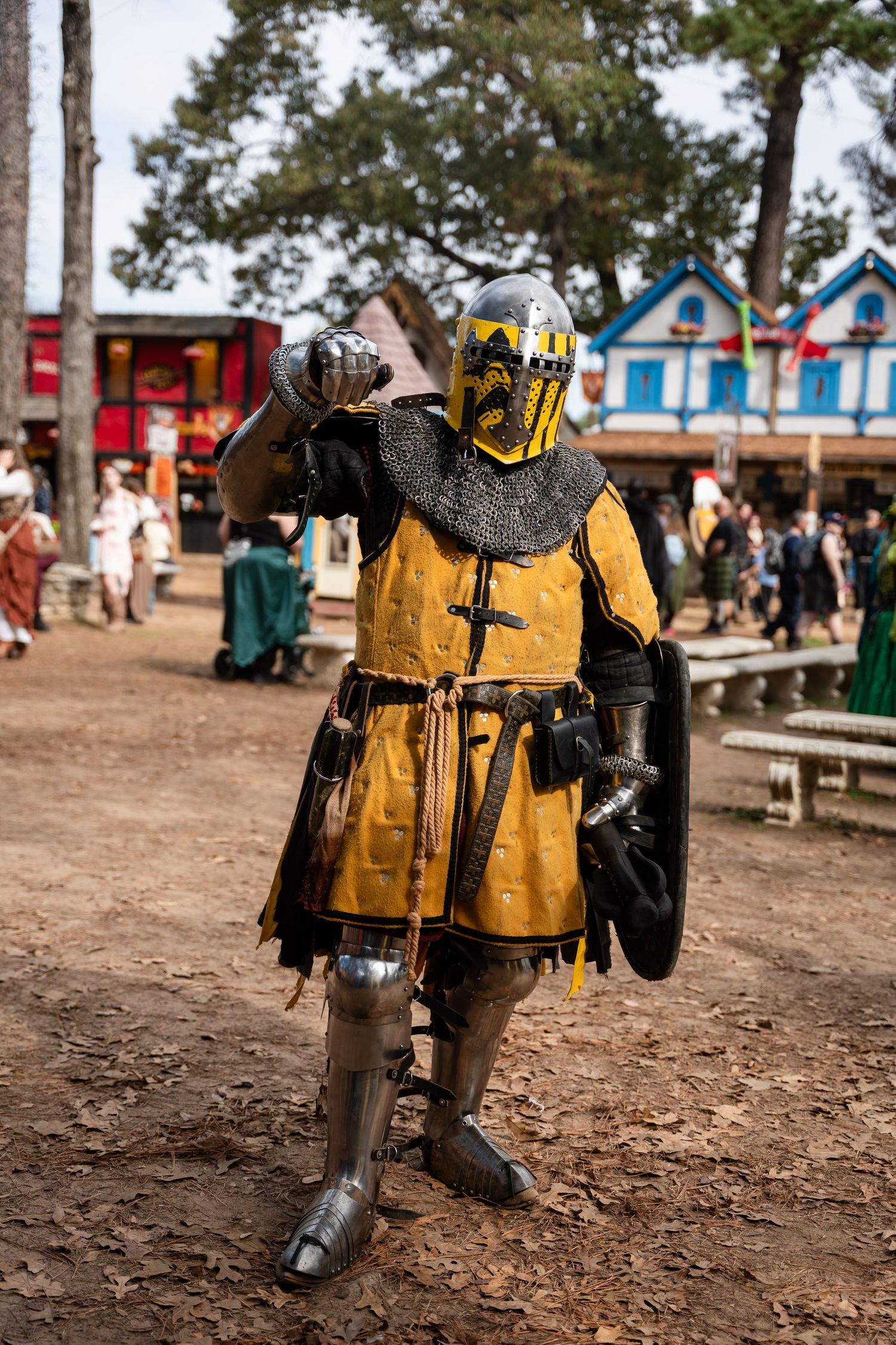 Medieval knight in yellow and black armor with sword and shield stands on dirt ground at renaissance festival.