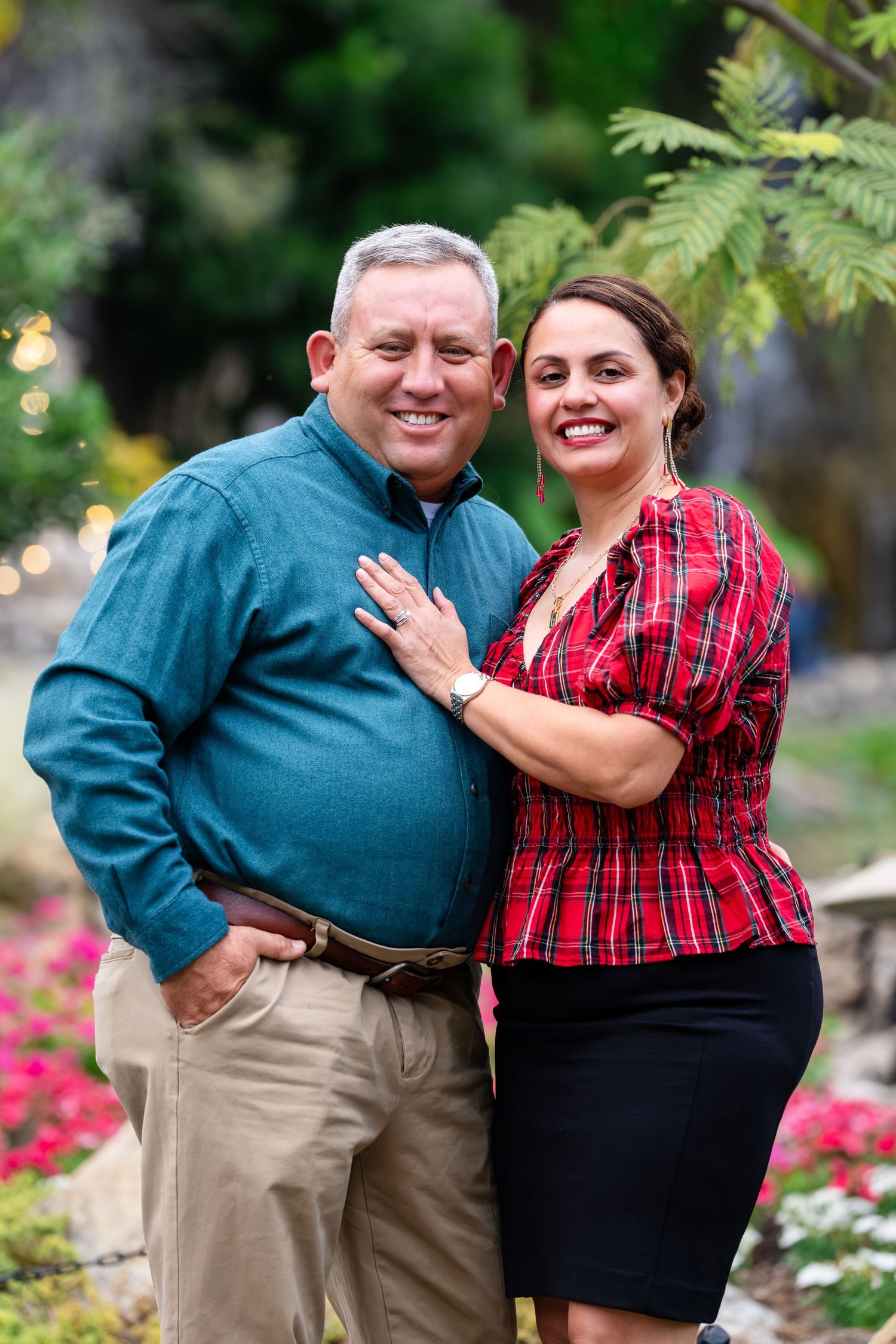A couple wearing coordinating plaid and dark clothing embraces while standing in a garden with blooming flowers.