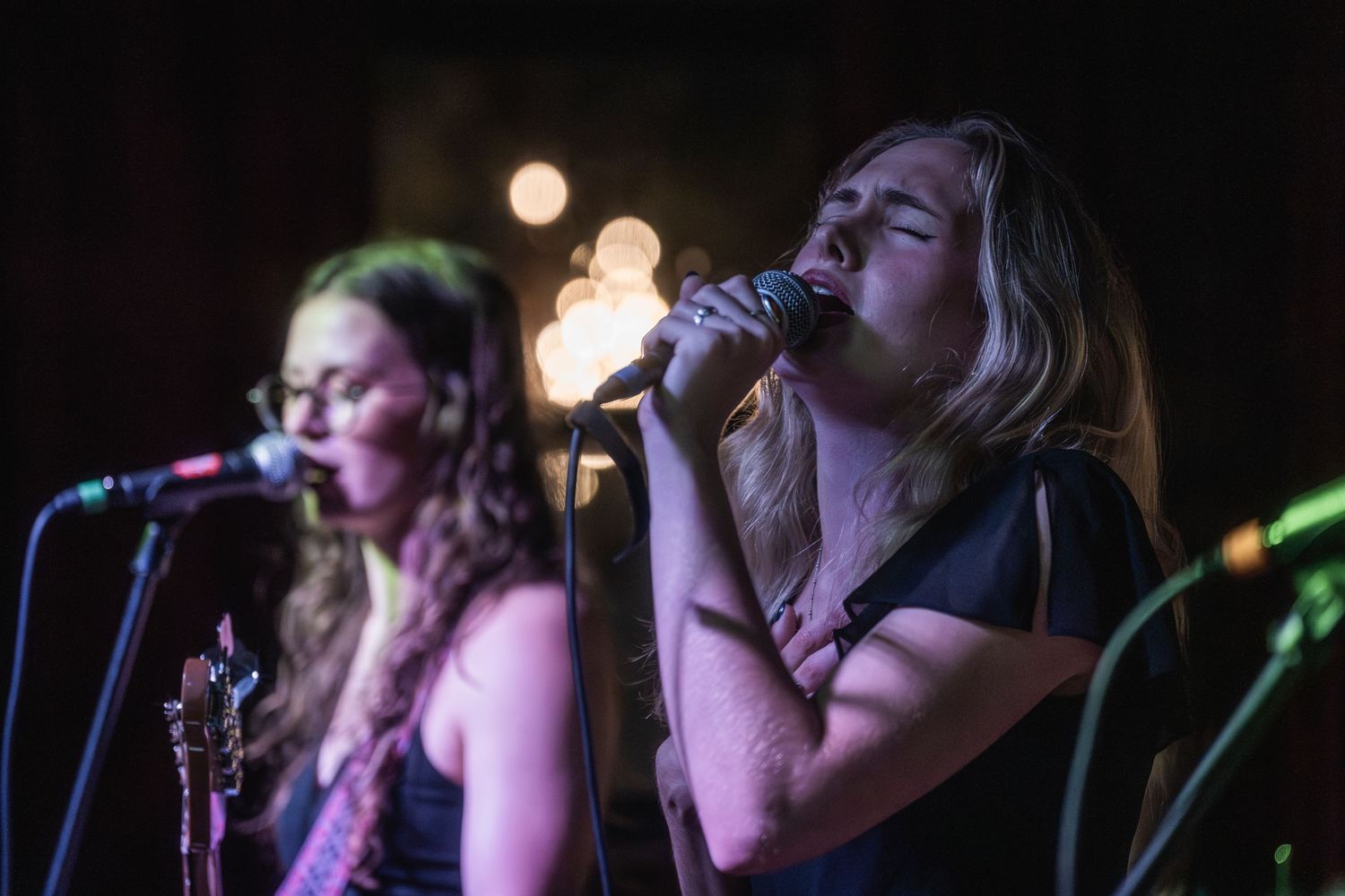 Musicians perform on stage at a dimly lit concert venue with dramatic lighting effects in the background.