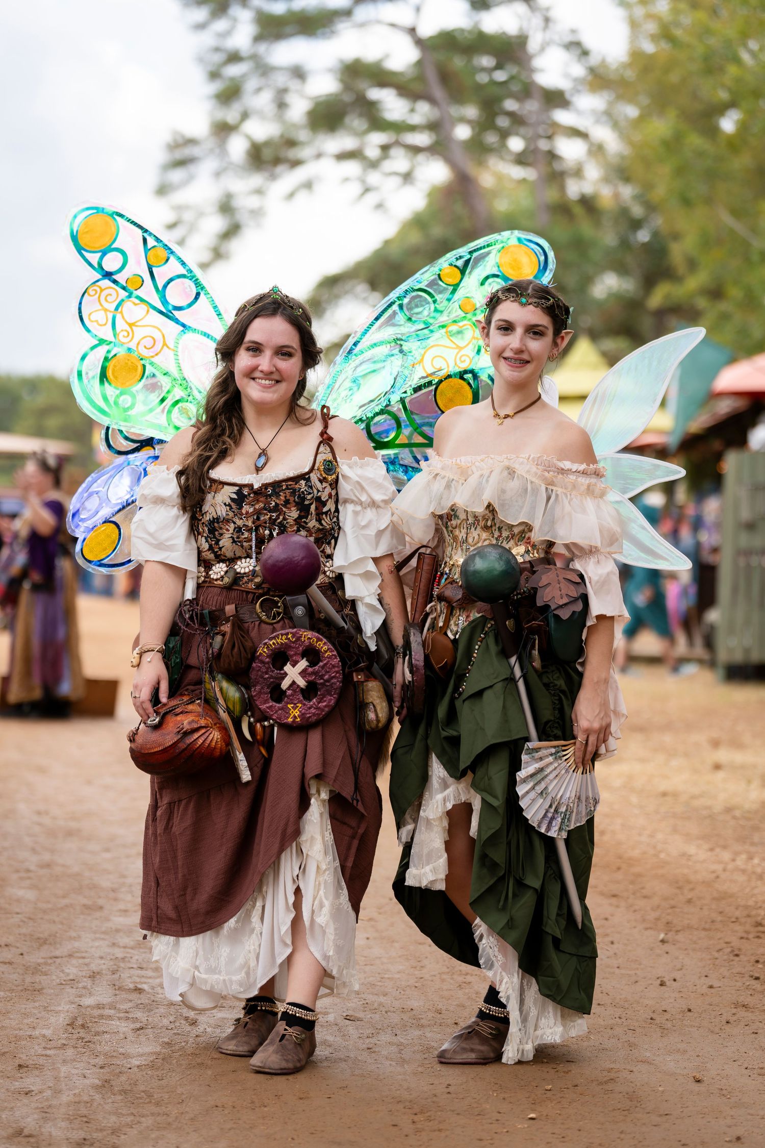 Two people in medieval renaissance fair costumes pose with colorful fairy wings at an outdoor festival.