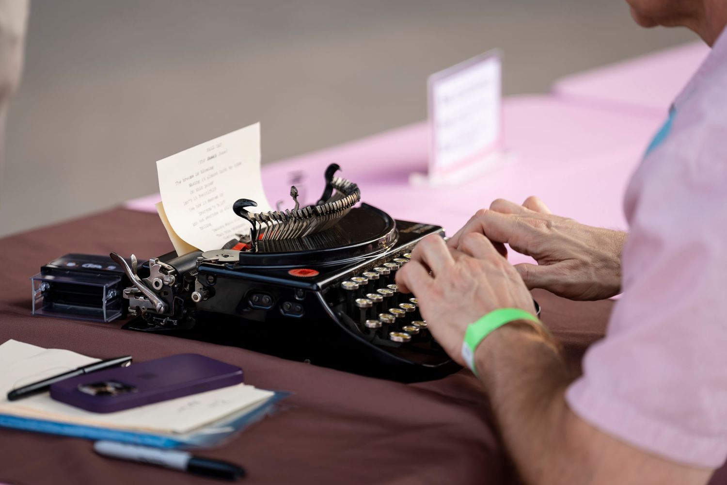Hands typing on a vintage manual typewriter keyboard with paper documents on a desk in soft pink lighting.