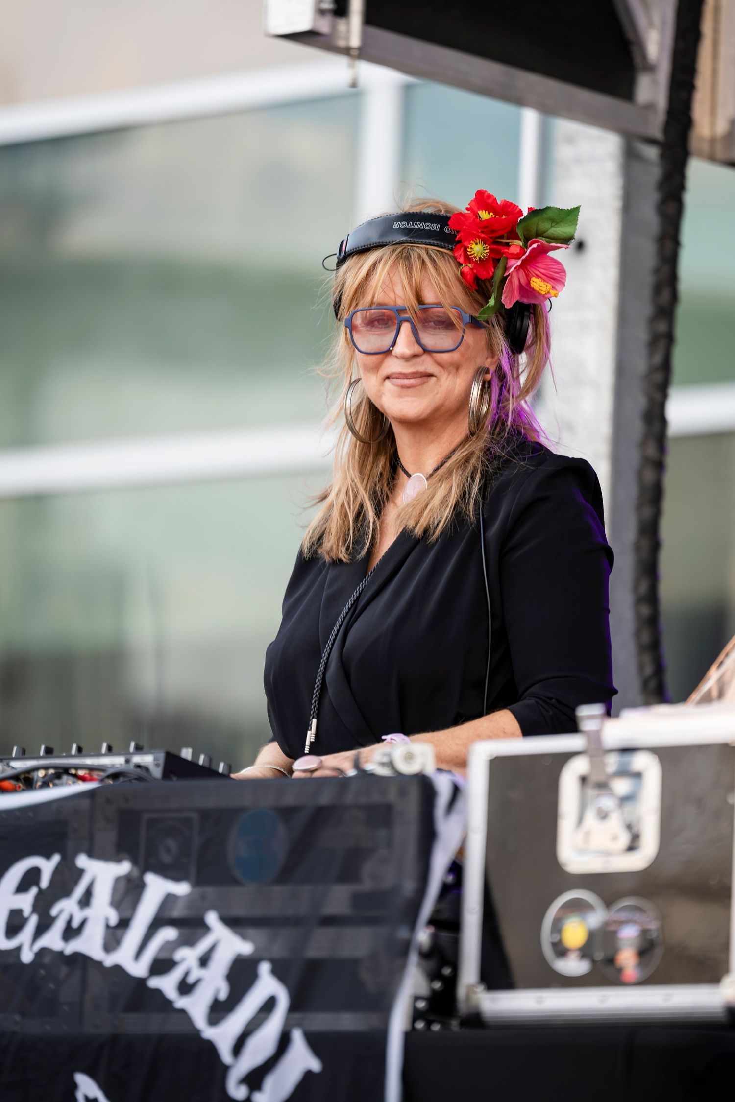 A DJ wearing glasses and black clothing performs behind decks at an outdoor event with red floral accessories.