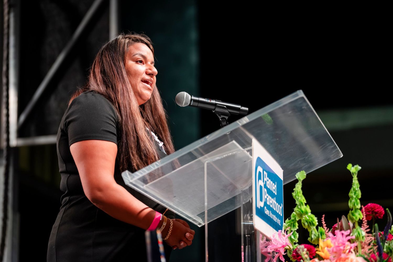A speaker stands at a clear podium with pink and green floral decorations during an outdoor event.