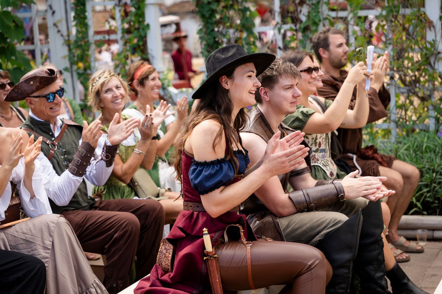 Wedding guests laugh and clap while seated outdoors during a joyful celebration in a garden setting.