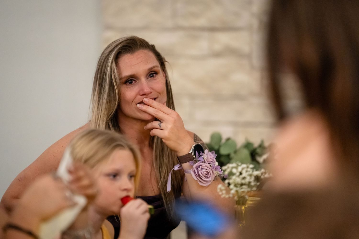 Emotional woman with blonde hair covering her mouth at a wedding reception, with a child beside her.