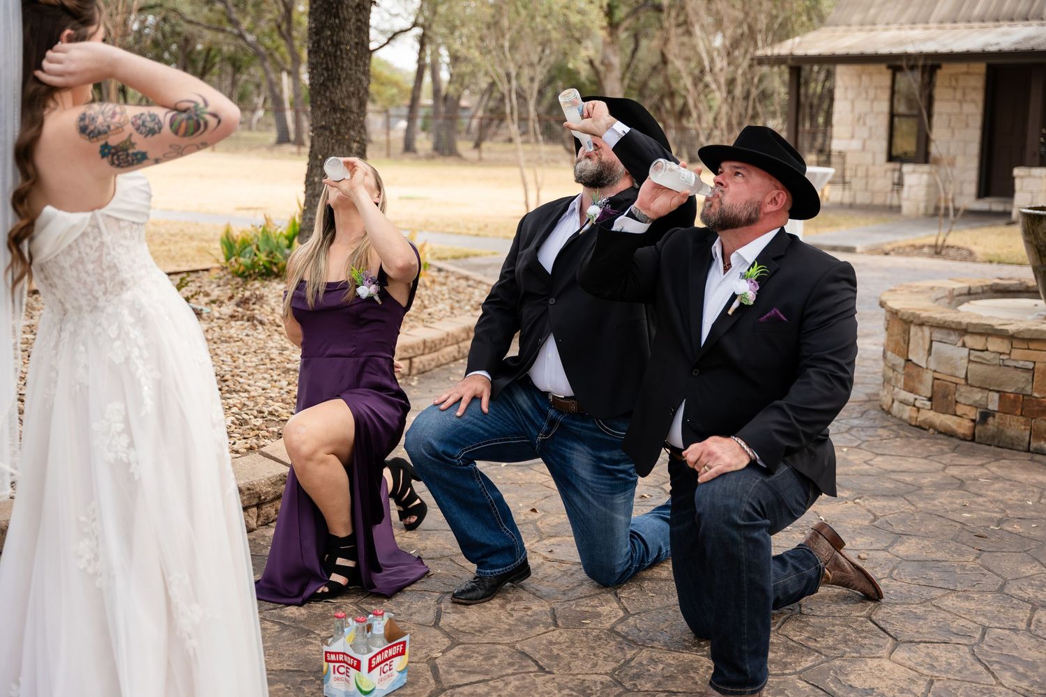 Wedding guests kneel and drink from cans outdoors while bride in white lace dress and woman in purple dress look on.
