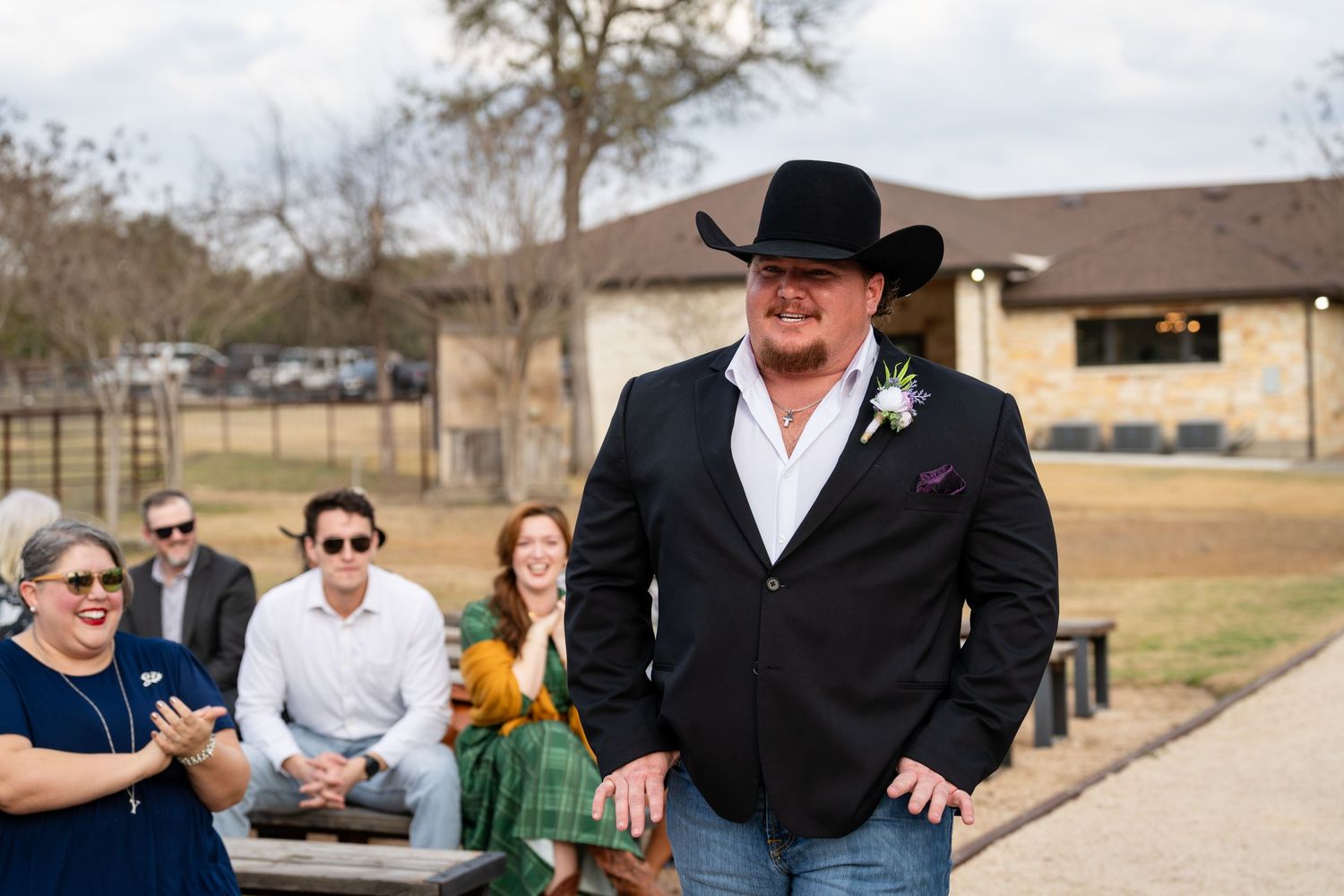 Groom in black blazer, cowboy hat, and jeans walks down outdoor wedding aisle with guests seated beside him.
