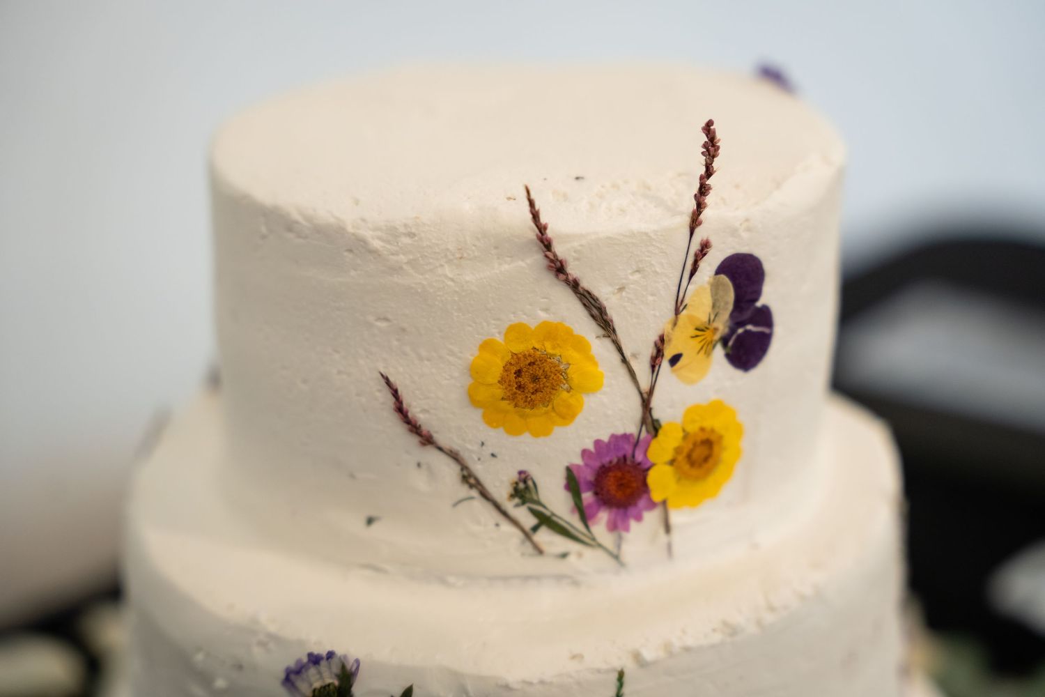 White tiered wedding cake decorated with pressed wildflowers including yellow, purple, and pink blooms.